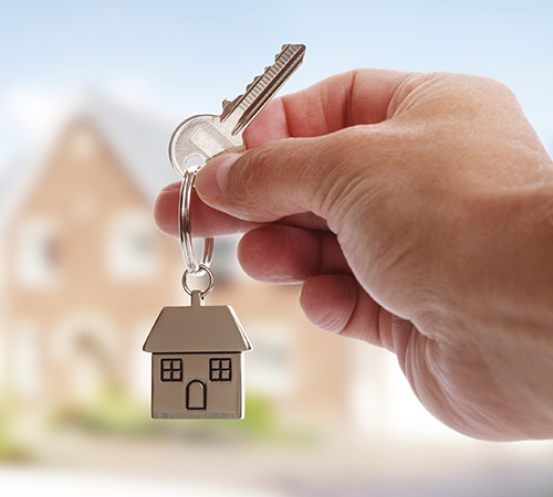 Hand holding house keys on a house-shaped keychain in front of a home